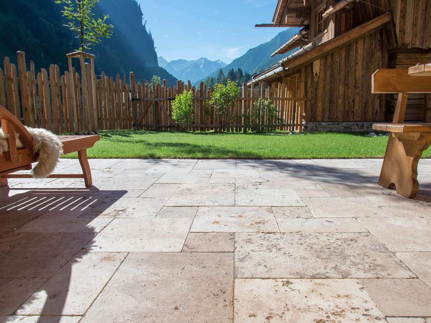 Sanfte, warme Beigetöne bei Sonnenlicht (Kundenprojekt) Terrassenboden mit Travertin Rustic und Holzmöbeln, im Hintergrund eine Rasenfläche und Ausblick auf die Berge