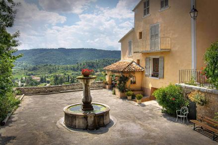 Ein Brunnen auf einem Platz neben einem Landhaus, im Hintergrund Berge