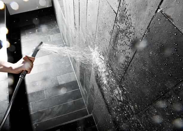 Hand hält eine Duschbrause gegen eine Wand mit dunklen Steinfliesen